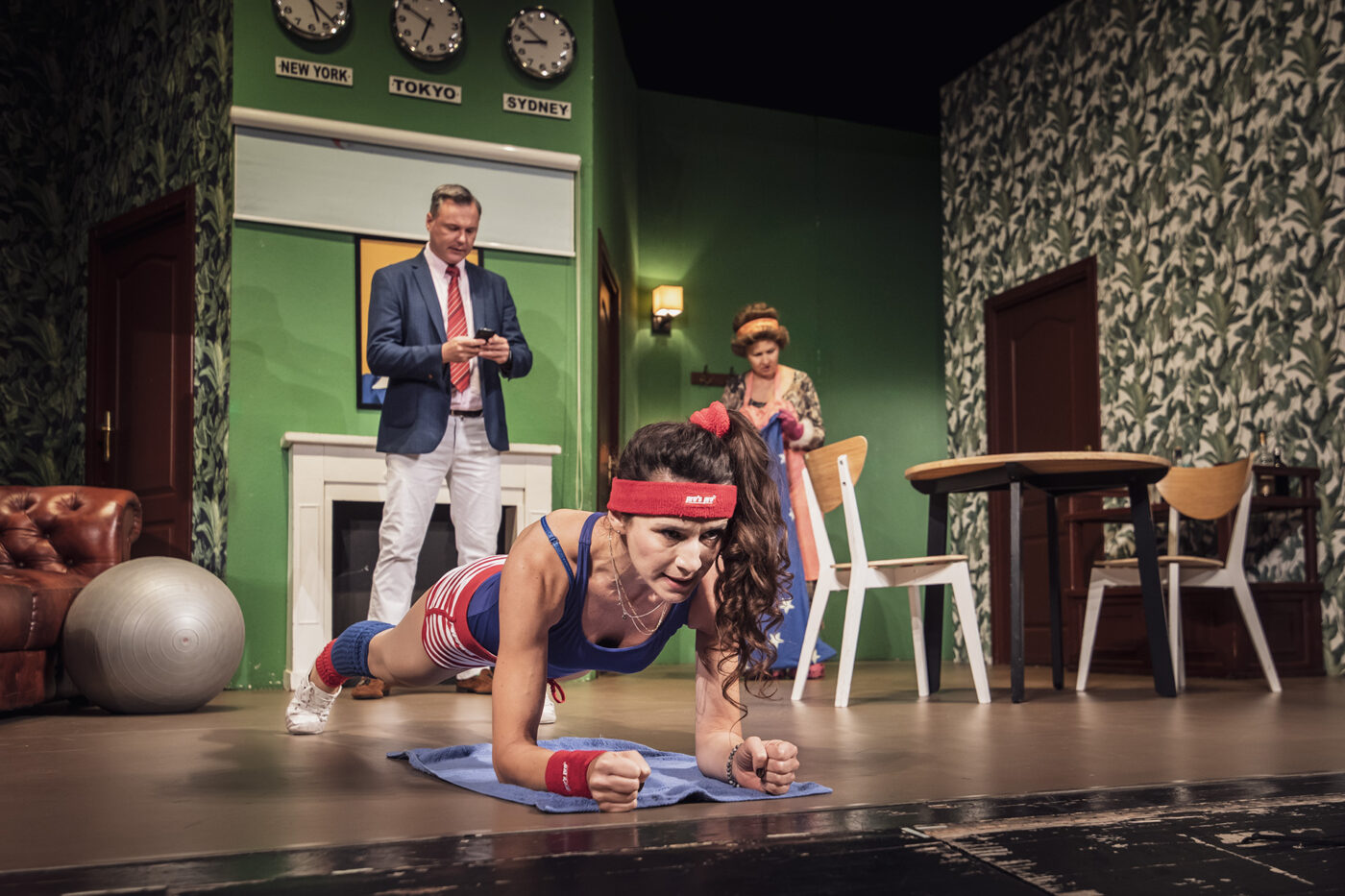 Aktorka w stroju sportowym wykonuje plank na scenie, za nią inni bohaterowie.