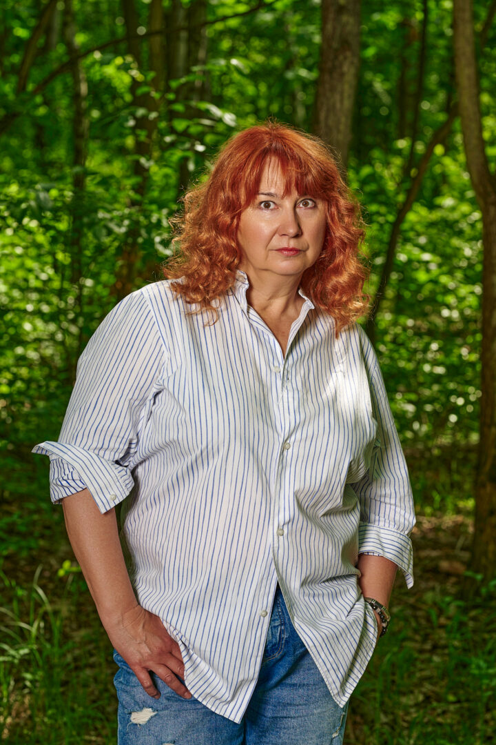 Kobieta o rudych włosach do ramion z grzywką na tle lasu. Trzyma ręce w kieszeniach jeansów. Ma ubraną niałą koszulę w prążki.