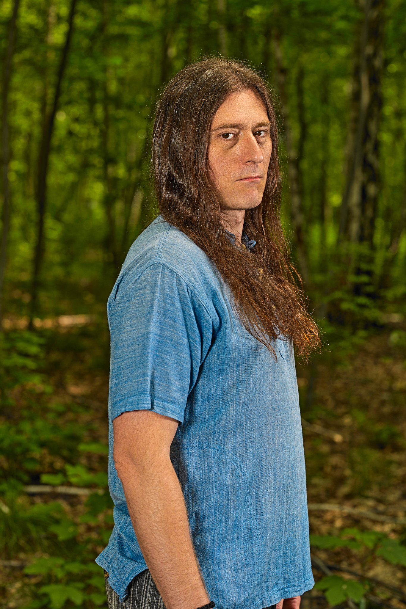 Mężczyzna na tle lasu. Aktor w długich, brązowych włosach. Ubrany w lnianą, niebieską koszulę z krótkim rękawem.