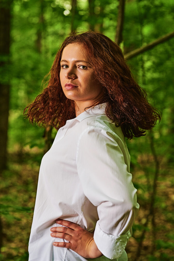 Młoda kobieta na tle lasu. Aktorka o cienych, kręconych włosach sięgających do ramion spogląda zza lewe ramię. Jest ubrana w białą koszulę.
