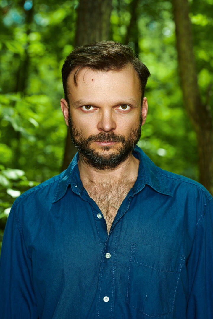 Mężczyzna na tle lasu. Aktor w krótkich, brązowych włosach z brodą. Ma ubraną niebieską koszulę z kołnierzykiem.