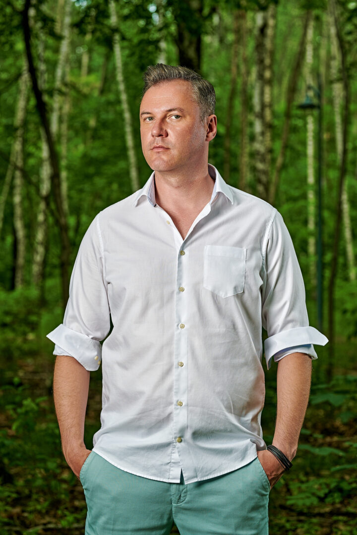 Mężczyzna na tle lasu. Siwiejący aktor ubrany w białą koszulę. Trzyma ręce w kieszeniach jasnozielonych spodni.