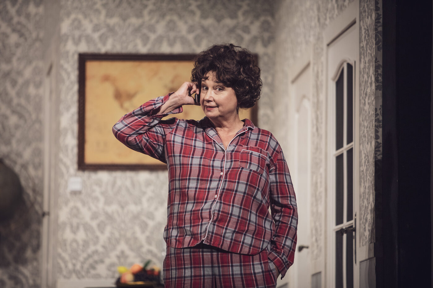 Kobieta w piżamie w czerwono-szarą kratę stoi z telefonem przy uchu, patrząc z figlarnym uśmiechem w bok. Jej druga ręka spoczywa w kieszeni spodni, a w tle widać mapę świata i drewniany sekretarzyk z owocami na górze.