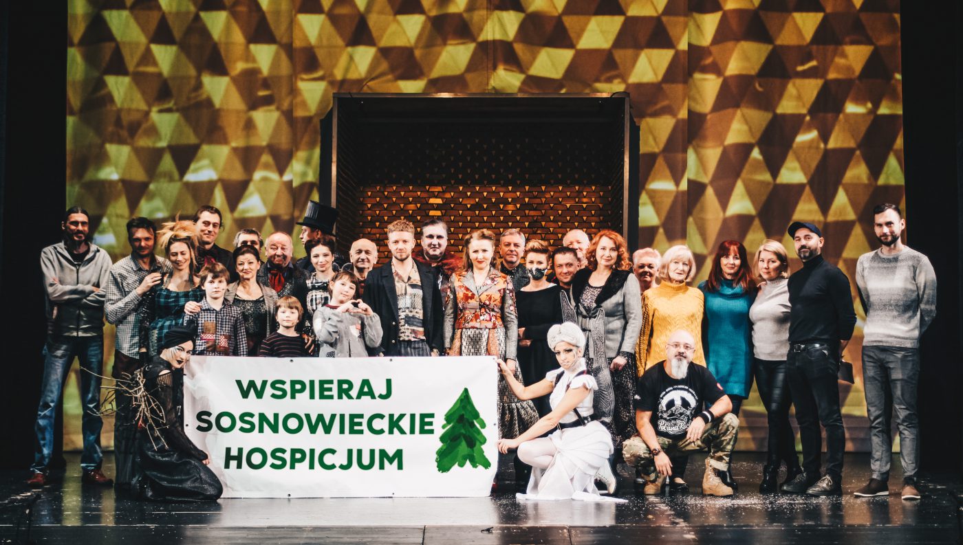 Grupa aktorów i pracowników Teatru na scenie. Na tle złotej scenografii trzyma banner z napisem Wspieraj sosnowieckie hospicjum.
