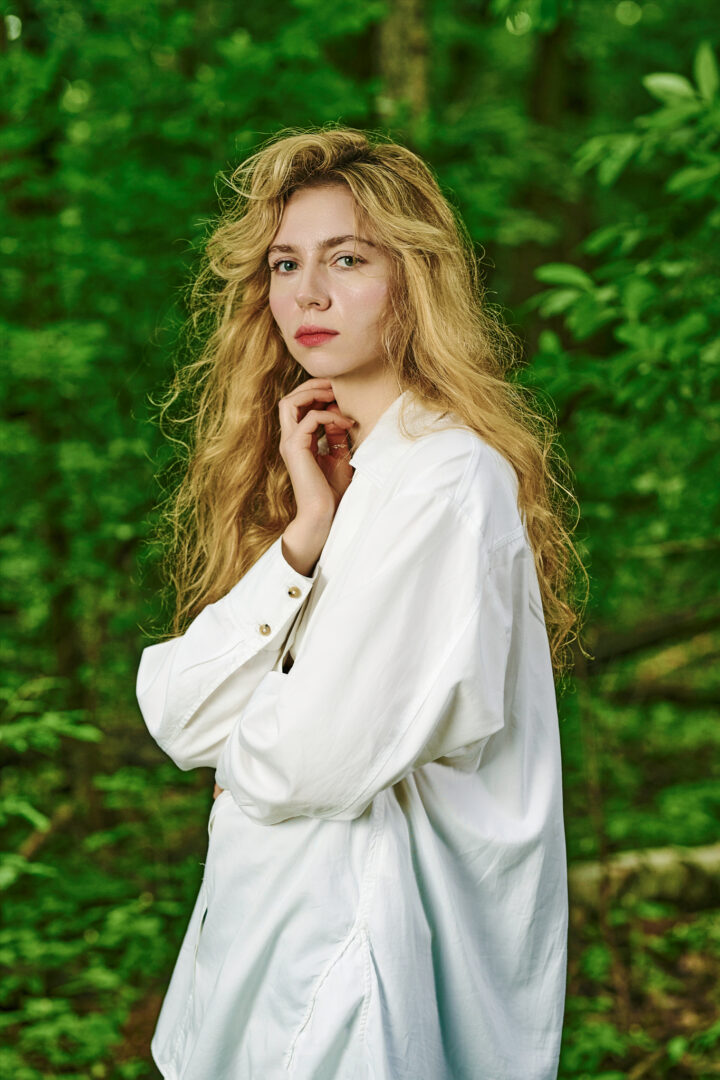 Kobieta na tle lasu. Młoda kobieta o długich blond włosach. Ma ubraną dużą, białą koszulę.