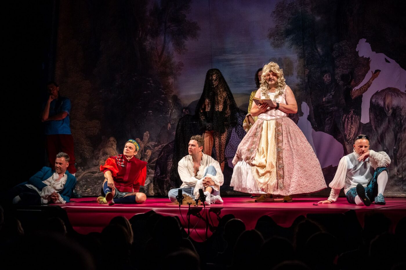Scena z Poskromienia złośnicy. Mężczyzna w stronu kobiety (grający rolę Katarzyny) stoi na scenie w blond peruce i odczytuje list. Reszta bohatwrów spektaklu siedzi na scenie z różowej wykładziny.