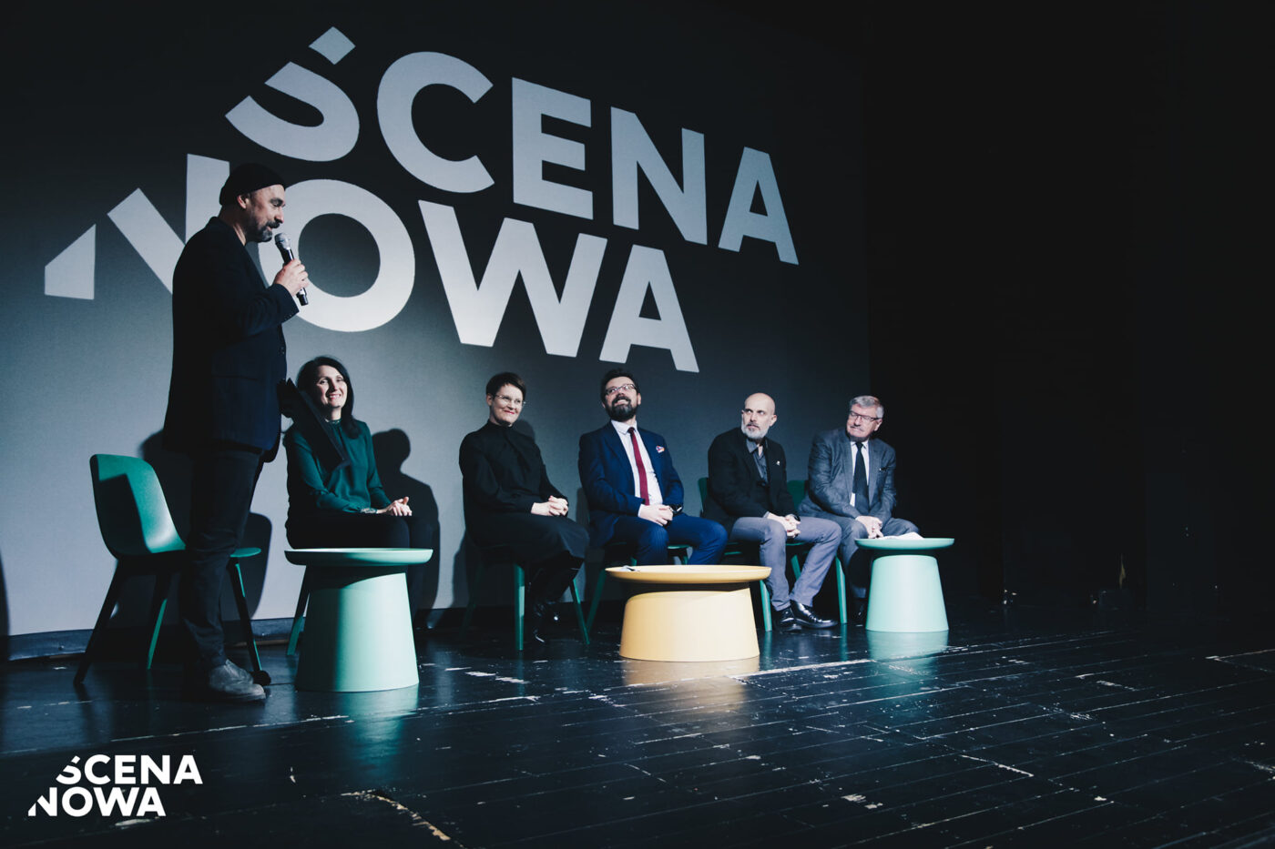 Zastępca dyrektora do spraw artystycznych przemawiający do mikrofonu na scenie. Pięć osób siedzących na scenie za stolikami, patrzą się na przemawiającego. W tle wyświetlane logo SCENA NOWA.