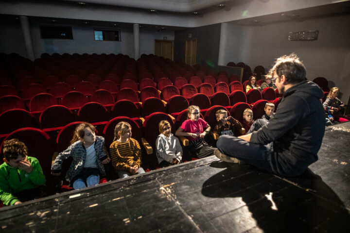 Aktor z dzećmi na widowni teatralnej. Aktor siedzi po turecku na scenie. Dzieci siedzą na czerwonych fotelach widowni.