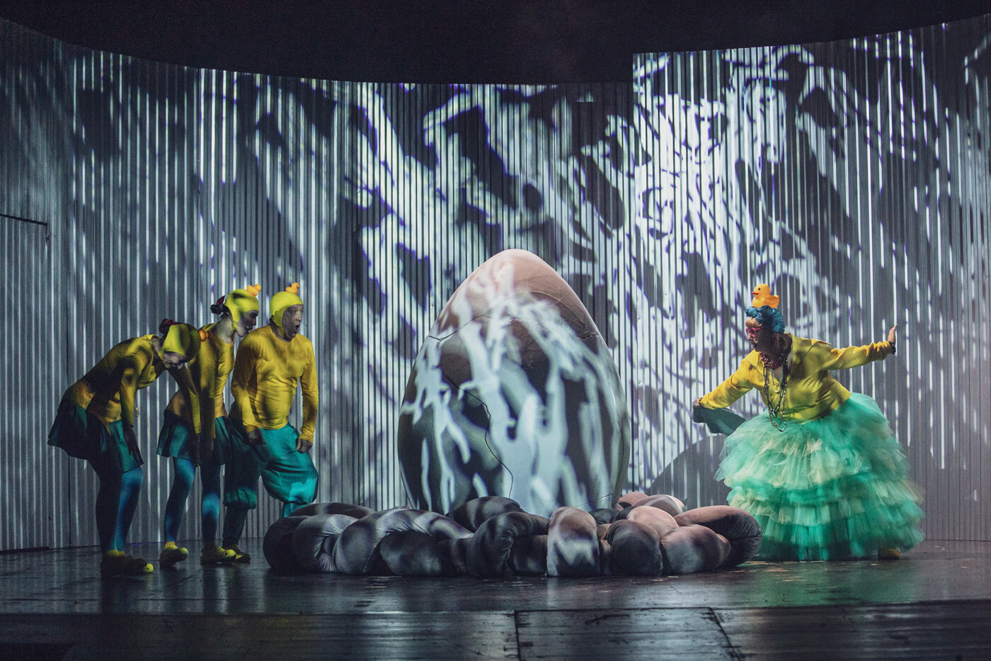 Na scenie stoi wielkie jajko otoczone gniazdem z szarych poduch, podświetlone ruchomymi projekcjami. Z lewej strony przyglądają się mu trzy postacie w żółto-zielonych kostiumach kaczątek z czapkami w kształcie kaczych głów. Z prawej kolorowo ubrana aktorka w turkusowej tiulowej spódnicy i żółtej bluzie gestykuluje z przejęciem.